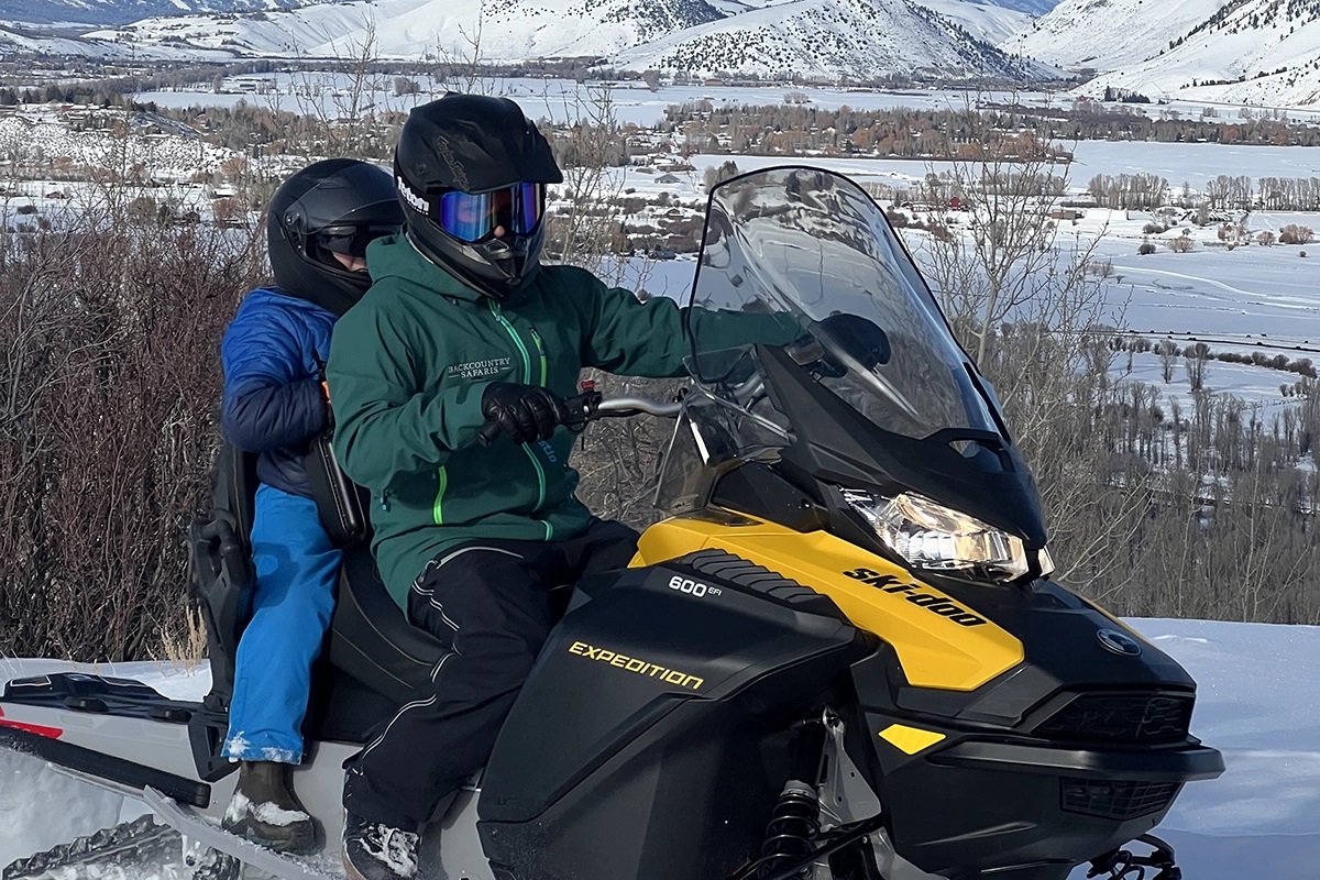 a person with a child on a snowmobile on a mountain with the valley in the background